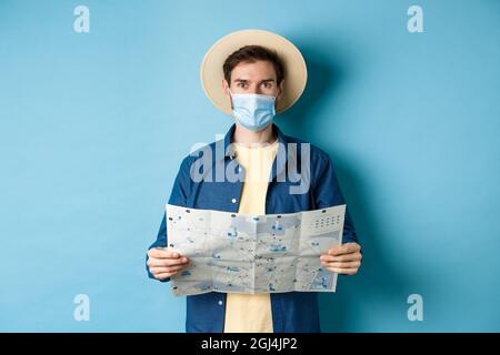 Covid-19, Pandemie- und Reisekonzept. Glücklicher Tourist im Sommerurlaub trägt medizinische Maske und Strohhut, hält Straßenkarte, auf blau stehen Stockfoto