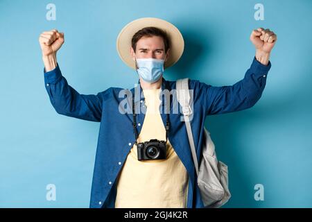 Covid-19 und Sommerferien Konzept. Fröhlicher, lächelnder Tourist, der seinen Urlaub feiert, Gesichtsmaske trägt, mit Freude Ja schreit und die Hände hochhebt Stockfoto