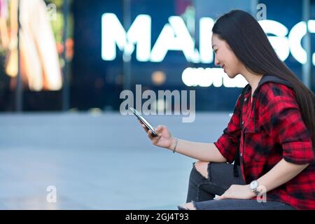Seitenansicht einer attraktiven Hipster-Frau in trendigem Outfit, die ihr Mobiltelefon nutzt und ihre Freunde darauf wartet, gemeinsam im Einkaufszentrum einkaufen zu gehen Stockfoto