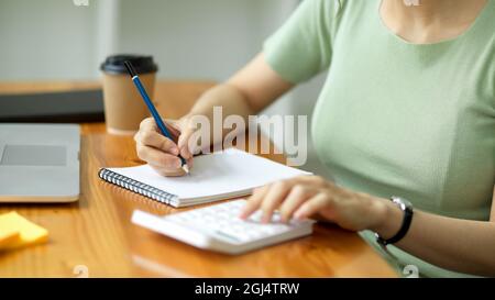 Junge Frau, die Notizen macht und mit einem Rechner am Schreibtisch berechnet, Haushaltsausgaben, Rechnungen zahlt, Steuern berechnet, Geschäftsbudget auswertet, ta Stockfoto