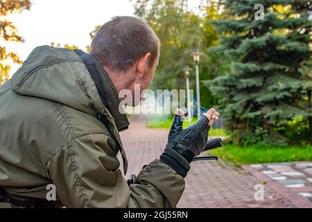 Die Hand eines nachdenklichen Mannes hält eine rauchende Zigarette, die auf der Straße der Stadtallee des Parks steht, ein Blick von hinten, hinten Stockfoto