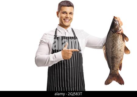 Junger glücklicher Mann, der eine Schürze trägt, einen großen Karpfenfisch hält und isoliert auf weißen Hintergrund zeigt Stockfoto