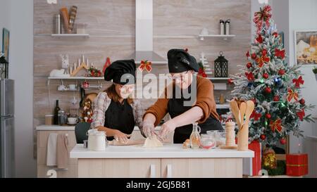 Großeltern genießen Winterferien, während Enkelkind mit Schürze Zubereitung von hausgemachten Cookies Dessert Herstellung Teig mit Nudelholz. Kulinarische Küche mit weihnachtsschmuck geschmückt Stockfoto