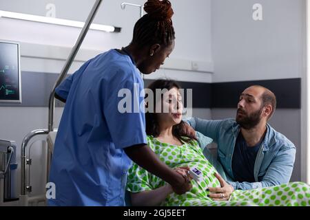 Eine Schwangere, die unter Schmerzen leidet, erhält medizinische Hilfe von einer schwarzen Krankenschwester, während sie im Krankenbett sitzt. Junger Mann, der die Ehefrau bei der Vorbereitung auf die Geburt unterstützt. Multiethnische Menschen mit Kindergeburten Stockfoto