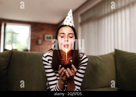 Geburtstagskind Brünette in Papphut sitzt auf der Couch oder dem Sofa und bläst brennende Kerze auf dem Cupcake aus. Frau hält Geburtstagskuchen in den Händen. C Stockfoto