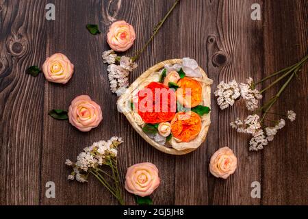 Schokolade blüht in einem Korb in Form eines Herzens. Rosen, Pfingstrosen auf braunem Holzhintergrund. Stockfoto