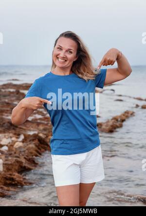 Lächelnde Frau mittleren Alters trägt ein blaues T-Shirt, zeigt darauf und steht bei Sonnenuntergang auf der Klippe. T-Shirt-Modell Stockfoto