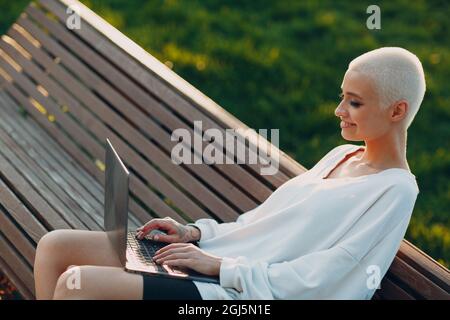 Porträt einer jungen, lächelnden, tausendjährigen europäischen Kurzhaarigen, die auf der Bank auf einer grünen Wiese im Park einen Laptop benutzt. Schöne glücklich blonde Mädchen im Freien. Stockfoto