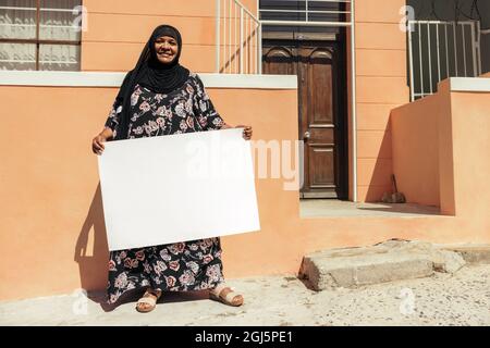 Lächelnde muslimische Frau mit einem leeren Plakat und Blick auf die Kamera, während sie tagsüber vor ihrem Haus steht. Selbstbewusste Frau mittleren Alters Stockfoto