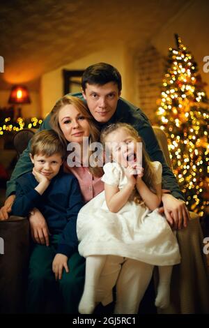 Junges schönes Paar mit zwei Kindern, das in einem Stuhl in einem weihnachtlichen Interieur mit einem weihnachtsbaum und Girlanden posiert. Selektiver Weichfokus, Filmkorneffekt Stockfoto