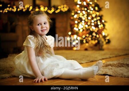 Porträt eines blonden niedlichen Mädchen 3-5 Jahre alt sitzt auf dem Boden vor dem Hintergrund eines festlichen Weihnachtsbaum. Selektiver Weichfokus, Filmkorneffekt Stockfoto
