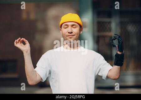 Behinderter europäischer Mann mit künstlichem Handglied, der im Freien meditiert. Hübscher junger Kerl trägt T-Shirt und Hut genießt sonnigen Tag. Konzept der psychischen Gesundheit. Moderne Medizintechnik heute Stockfoto