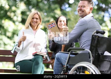 Behinderter Mann im Rollstuhl studiert Englisch mit Freundinnen im Park. Stockfoto