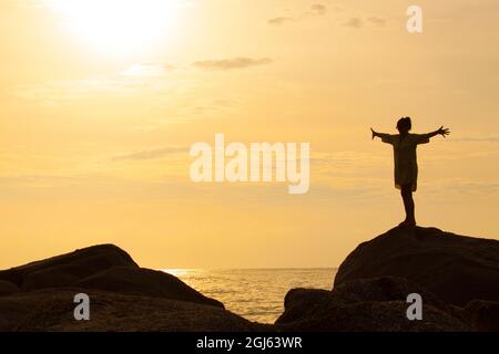 Thailand, Ko Samui. Silhouette einer Frau, die mit ausgestreckten Armen auf einem Felsen in der Nähe des Ozeans steht. Stockfoto