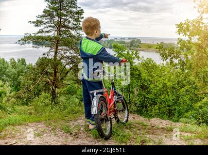 Das Kind mit dem Fahrrad steht hoch auf einem Hügel mit Blick auf den Fluss. Stockfoto