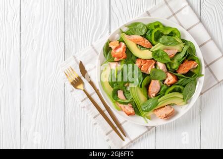 Lachs Spinat Avocado und grüner Pfeffer Salat auf einem weißen Teller mit goldener Gabel und Messer auf einem weißen Holztisch, mediterrane gesunde Ernährung, horizonta Stockfoto