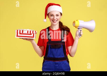 Lächelnde Kurierin hält Geschenk-Box und Megaphon in den Händen, schaut auf die Kamera, kündigt Anzeige, trägt blauen Overalls und weihnachtsmann Hut. Innen Stockfoto