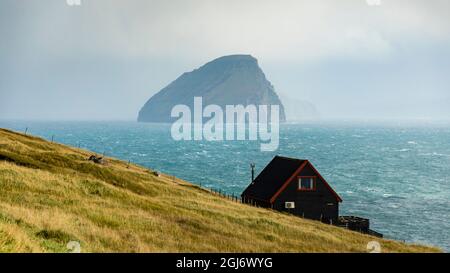 Europa, Färöer. Blick auf das traditionelle Haus auf einer Klippe am Meer auf der Insel Streymoy, mit der Insel Kuttur im Hintergrund. Stockfoto