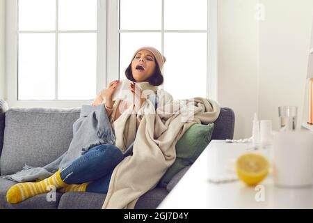 Kranke junge Frau, die an Erkältung oder Grippe leidet, zu Hause auf der Couch sitzt und niest Stockfoto