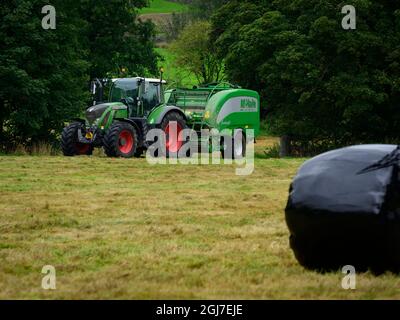 Heu- oder Silageherstellung (Landwirt im landwirtschaftlichen Traktor bei der Arbeit auf dem Feld, Ballenpresse ziehen, trockenes Gras sammeln und runden schwarzen Ballen verpackt - Yorkshire England, Großbritannien. Stockfoto