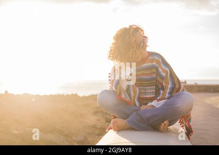 Wunderschöne Hipster-Frau, die auf einem Betondamm sitzt und die Aussicht mit der Meereslandschaft im Hintergrund bewundert. Silhouette einer Frau, die ihren Urlaub am Meer genießt Stockfoto