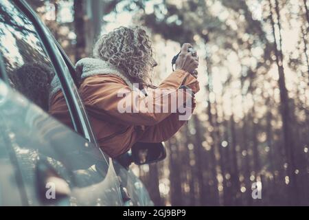 Frau in warmer Jacke, die sich aus dem Autofenster lehnt und mit einer dslr-Kamera entlang des Waldes erkundet und fotografiert. Frau mit lockigen Haaren und warmer Jacke t Stockfoto