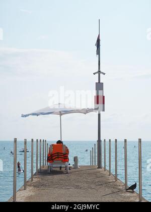 Rettungsschwimmer sitzt an einem sonnigen Sommertag in einer Sonnenliege am Pier unter einem Sonnenschirm am Meer Stockfoto