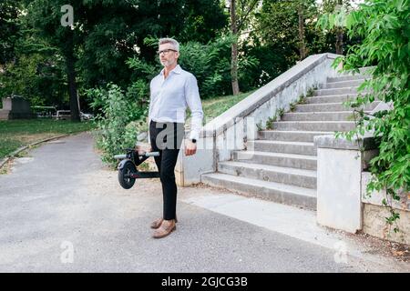 Senior Geschäftsmann hält Elektroroller in der Hand, während er in der Stadt draußen geht Stockfoto