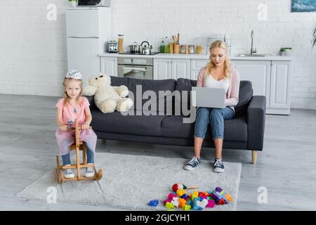 Mädchen im Kostüm der Prinzessin Reiten Schaukelpferd in der Nähe Mutter arbeitet auf dem Laptop auf dem Sofa Stockfoto