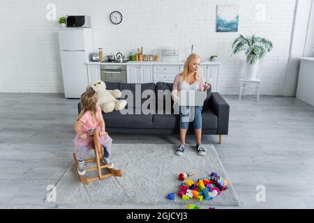 Mädchen reiten Schaukelpferd im Kostüm der Prinzessin in der Nähe Mama arbeiten auf Laptop auf dem Sofa Stockfoto