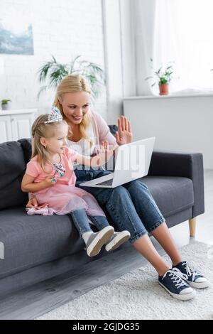 Fröhliche Frau und Mädchen in der Spielzeugkrone winken die Hände während eines Videoanrufs auf dem Laptop Stockfoto