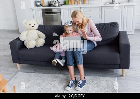 Glückliche Frau mit Tochter in Spielzeug Krone winkende Hände während Videoanruf zu Hause Stockfoto