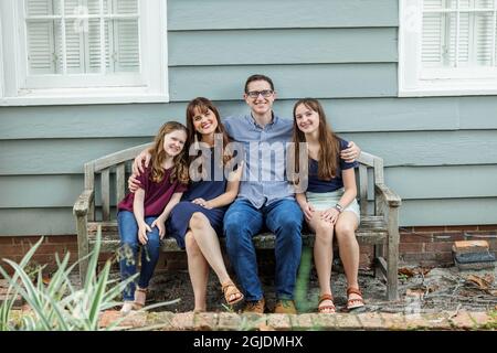 Eine vierköpfige Familie mit Mutter, Vater und zwei Töchtern, die auf einer Bank vor einem blauen Häuschen sitzen Stockfoto