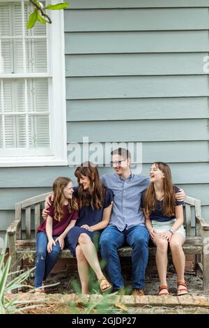 Eine vierköpfige Familie mit Mutter, Vater und zwei Töchtern, die auf einer Bank vor einem blauen Häuschen sitzen Stockfoto
