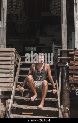 Auf der Holztreppe eines gemütlichen Café im asiatischen Stil sitzt ein junger, hübscher Kaukasier mit einem Ärmel-Tattoo. Mit Blick auf das Meer. Stockfoto