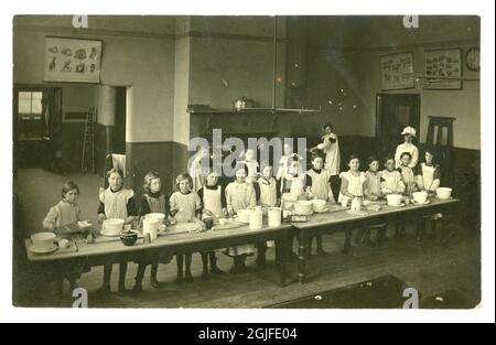 Original Postkarte aus dem frühen 20. Jahrhundert, Mädchen im Kochkurs, die an langen Tischen arbeiteten, Gemüse-, Fleisch- und Viehkarten an den Wänden, erstaunliche Backöfen, Brot und Scones backen - London, England, Großbritannien um 1903 Stockfoto