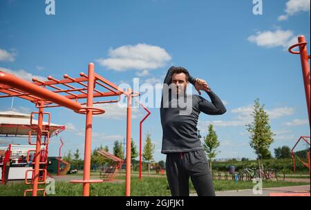 Schöner junger Erwachsener, 40 Jahre alter sportlicher Mann aus Europa, der die Arme hinter seinem Rücken streckt, bevor er im Freien auf dem Sportplatz trainiert. Ausgereifte Sportsma Stockfoto