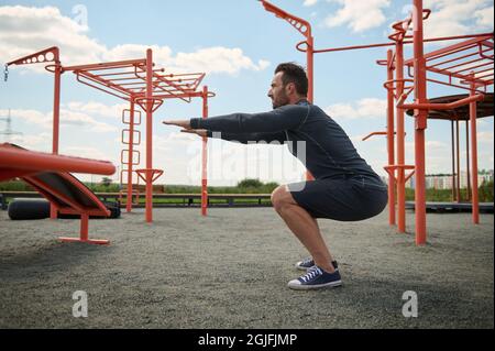 Aktiver Athlet in sportlicher Uniform, der beim Training auf einem Sportplatz im Freien Körpergewicht hockt. Junger bärtiger Mann, der im Sommer Sport macht Stockfoto