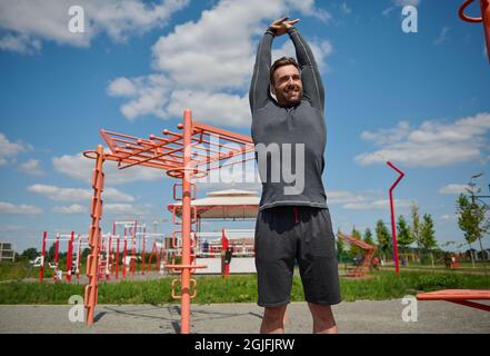 Sportlicher kaukasischer Europäischer Mann, der draußen arbeitet. Glücklicher Sportler, der im Freien Fitness-Workout macht, seine Arme hochhebt und die Verlängerung durchführt Stockfoto