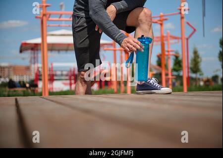Nicht erkennbarer männlicher Athlet Beine auf einem Sportplatz, Sportler nimmt eine Flasche mit Wasser, seine Körper während des Trainings im Freien rehydrieren. Nahaufnahme, Ernte Stockfoto