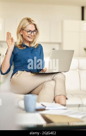 Attraktive reife Frau, die von zu Hause aus arbeitet und während eines Online-Videoanrufs mit einem Laptop auf einer Couch in einer modernen Wohnung winkt Stockfoto