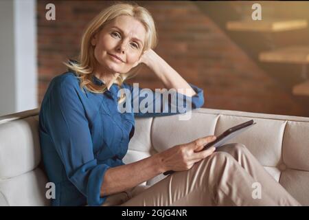 Eine ziemlich reife Frau, die die Kamera anschaut, während sie einen digitalen Tablet-pc hält und sich zu Hause auf der Couch in einer modernen Wohnung entspannt Stockfoto