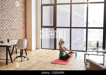 Die reife Sportlerin, die in Lotus sitzt, posiert auf einer Matte und benutzt einen Laptop, während sie zu Hause in einer modernen Wohnung Yoga praktiziert Stockfoto