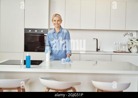 Fröhliche reife Hausfrau in Gummihandschuhen lächelt an der Kamera, während sie tagsüber zu Hause mit Waschmittel die Küche putzt Stockfoto