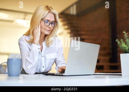 Beschäftigte reife Geschäftsfrau in Brille mit Laptop, während sie im Büro einige Schreibarbeiten machte Stockfoto