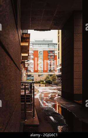 Blick auf helles Wohngebäude durch dunklen Bogen, regnerischer Tag, Pfützen auf Asphalt Stockfoto