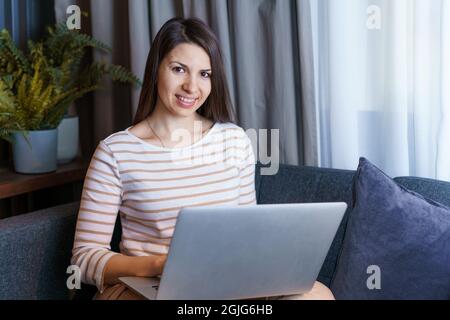 Junge Frau, die auf der Couch sitzt, am Laptop arbeitet, im Internet surft oder Nachrichten von zu Hause liest, glückliche Schülerin auf der Couch, Fernunterricht am Computer, Online-Recherche im Internet Stockfoto