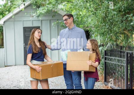 Ein Vater und seine beiden Töchter halten bewegliche Kisten auf einer Schottereinfahrt in der Nähe eines Lagergebäudes Stockfoto