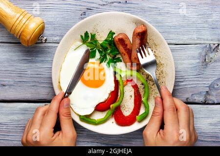 Sonnenseite oben. Spiegeleier mit Würstchen und Gemüse auf einem hellen Holztisch. Zwei Hände halten eine Gabel und ein Messer Stockfoto