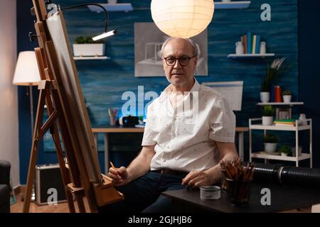 Porträt eines älteren kaukasischen Künstlers, der in einem Kunststudio sitzt. Älterer Mann mit künstlerischer Phantasie, der während der Arbeit an der Vasenzeichnung die Kamera anschaut. Gealterte Person mit Meisterwerk auf Leinwand Stockfoto